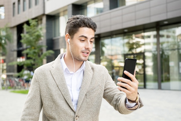 Giovane uomo d'affari contemporaneo di razza mista con auricolari che guarda lo schermo dello smartphone durante la comunicazione in ambiente urbano