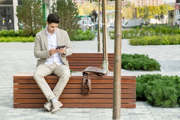 Giovane uomo d'affari contemporaneo in abbigliamento formale seduto su una panca di legno nel parco e scorrendo in tavoletta digitale in ambiente urbano