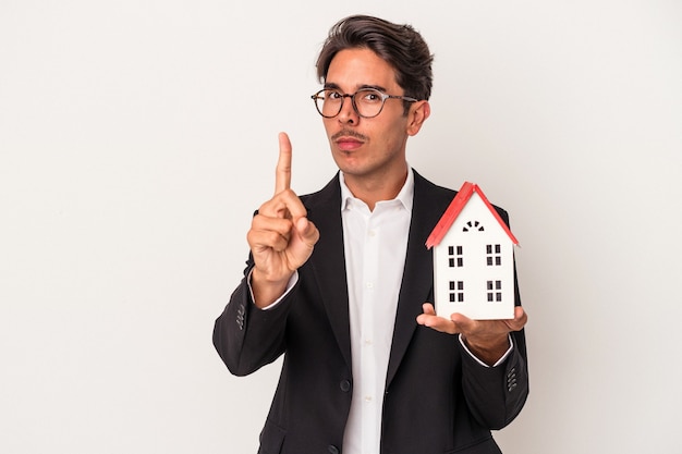 Giovane uomo d'affari di razza mista in possesso di una casa giocattolo isolata su sfondo bianco che mostra il numero uno con il dito.