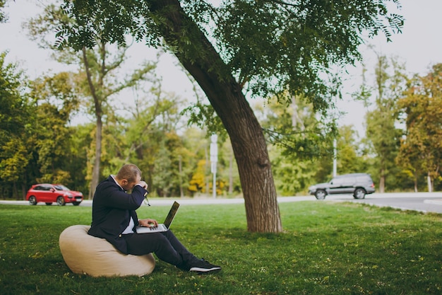 Giovane uomo d'affari di successo in camicia bianca, abito classico, occhiali. L'uomo si siede su un morbido pouf, lavorando su un computer portatile nel parco cittadino sul prato verde all'aperto sulla natura. Ufficio mobile, concetto di affari.