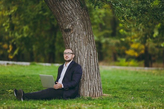 Giovane uomo d'affari di successo in camicia bianca, abito classico, occhiali. L'uomo si siede sul terreno erboso, lavora su un computer portatile nel parco cittadino sul prato verde all'aperto sulla natura. Ufficio mobile, concetto di affari.