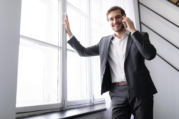 Giovane uomo d'affari di successo parla al telefono in una giacca alla finestra in ufficio