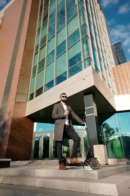 Giovane uomo d'affari elegante che beve caffè mantenendo una gamba su uno scooter elettrico contro un grande edificio per uffici contemporaneo