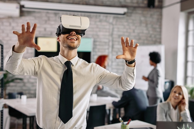Giovane uomo d'affari felice che testa il simulatore di realtà virtuale mentre lavora in ufficio Ci sono persone in background