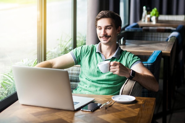 Giovane uomo d'affari felice in maglietta verde seduto e guardando il display del laptop con un sorriso che tiene e beve una tazza di caffè rilassandosi e godendosi il concetto di business tiro al coperto vicino alla finestra