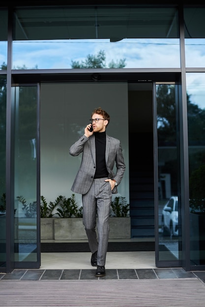 Giovane uomo d'affari fiducioso che indossa un abito elegante, occhiali che parlano al cellulare per strada