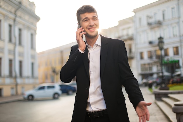 Giovane uomo d'affari gioioso in giacca nera e camicia bianca che parla felicemente al cellulare mentre guarda da parte con una bellissima vista sulla città sullo sfondo