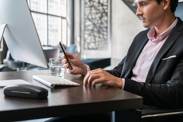 Giovane uomo d'affari guardando il telefono che indossa un abito nero seduto al tavolo con il computer Responsabile dell'ufficio con il telefono che lavora in un ufficio moderno con finestre di vetro sfondo sfocato concetto di lavoro