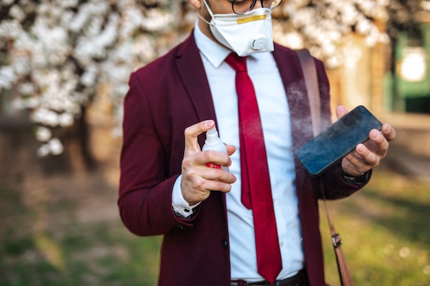 Giovane uomo d'affari in piedi nel parco, indossando una maschera protettiva e disinfettando lo smartphone con l'alcol.