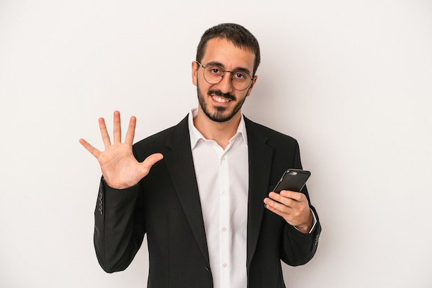 Giovane uomo d'affari in possesso di un telefono cellulare isolato su sfondo bianco sorridente allegro che mostra il numero cinque con le dita.