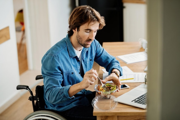Giovane uomo d'affari in sedia a rotelle che lavora a casa e mangia insalata in pausa pranzo