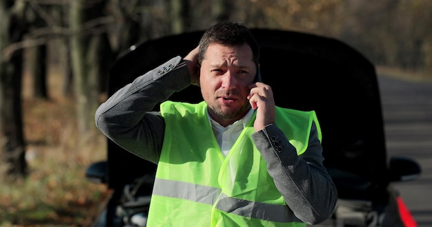 Giovane uomo d'affari in un giubbotto di sicurezza che chiede assistenza con la sua auto in panne sul ciglio della strada. L'uomo indifeso ha bisogno di aiuto e assistenza.