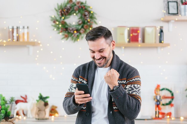 Giovane uomo d'affari libero professionista esulta vincendo felice vittoria legge grandi notizie dal telefono cellulare a casa durante le vacanze di Capodanno e Natale