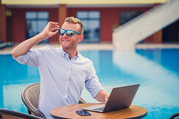 Giovane uomo d'affari maschio in occhiali da sole lavora a un computer portatile seduto a un tavolo vicino alla piscina Lavoro a distanza Freelancer