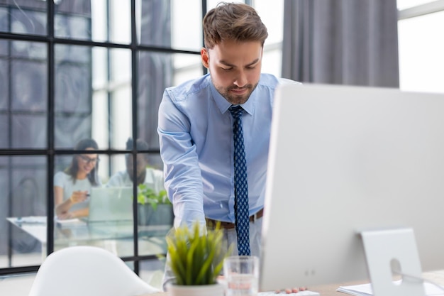 Giovane uomo d'affari moderno che analizza i dati utilizzando il computer mentre si lavora in ufficio con i colleghi sullo sfondo