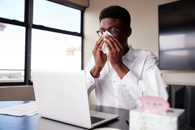 Giovane uomo d'affari nero seduto alla scrivania di un ufficio che soffia il naso in un tessuto