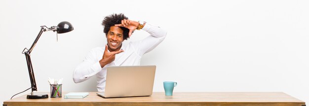 Giovane uomo d'affari nero sentirsi felice, amichevole e positivo, sorridendo e facendo un ritratto o una cornice con le mani su una scrivania