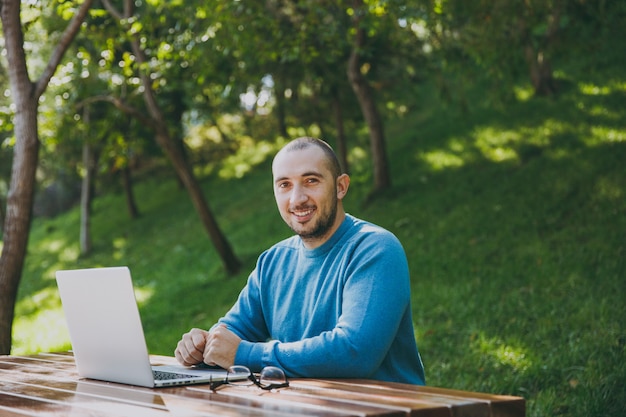 Giovane uomo d'affari o studente intelligente di successo con gli occhiali casual della camicia blu seduto al tavolo con il telefono cellulare nel parco cittadino utilizzando il computer portatile che lavora all'aperto sulla natura verde. Concetto di ufficio mobile.