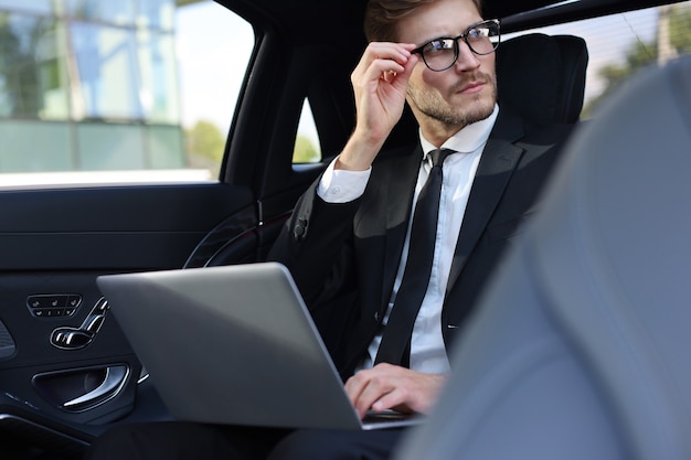 Giovane uomo d'affari premuroso che tiene la mano sugli occhiali mentre è seduto nell'auto lux e usa il suo laptop.