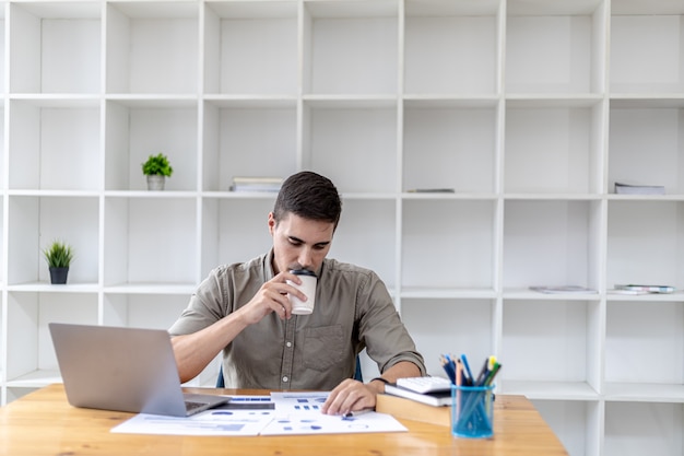 Giovane uomo d'affari seduto in ufficio con un laptop, facendo una pausa caffè calda mentre controlla i documenti, ha fondato una startup, è un giovane imprenditore che ha le competenze per gestire un'azienda in crescita.