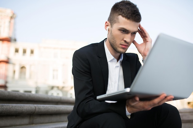 Giovane uomo d'affari serio nella classica giacca nera e camicia bianca con auricolari wireless che lavora premurosamente sul laptop mentre trascorre del tempo all'aperto