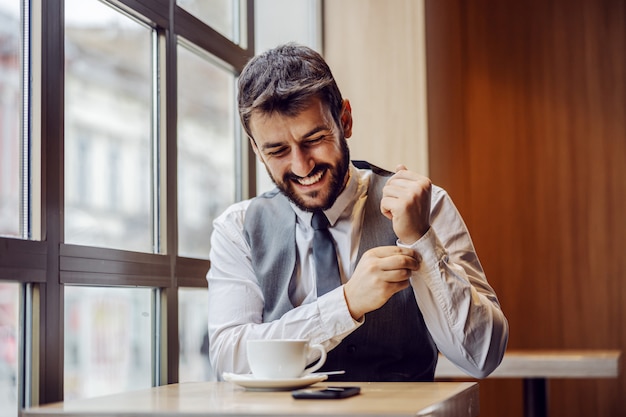 Giovane uomo d'affari sorridente che si siede nella caffetteria sulla pausa caffè e abbottona il gemello.