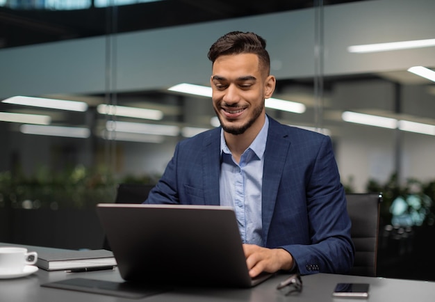 Giovane uomo d'affari sorridente del Medio Oriente seduto al tavolo di lavoro in un ufficio moderno che scrive al computer