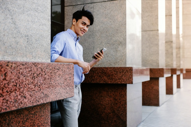 Giovane uomo d'affari sorridente di Asain che utilizza telefono cellulare nella città. Guardando la fotocamera