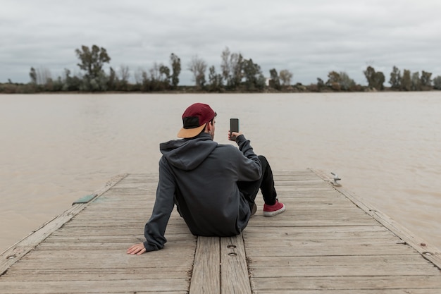 Giovane uomo da dietro che indossa un maglione grigio e un berretto rosso utilizzando uno smartphone e scattare foto seduto in un molo vicino a un fiume in una giornata nuvolosa