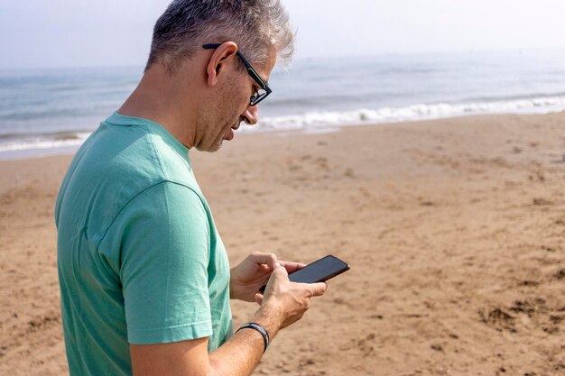 Giovane uomo dai capelli bianchi che guarda il suo cellulare mentre cammina sulla spiaggia