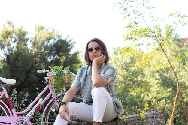 Giovane uomo dai capelli lunghi bello su occhiali da sole seduto su un tronco che guarda alla macchina fotografica con una bici rosa dietro