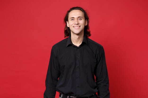 Giovane uomo dai capelli lunghi ricci alla moda bello in camicia nera che posa isolata sul ritratto rosso dello studio del fondo della parete. Persone sincere emozioni concetto di stile di vita. Mock up spazio di copia. Guardando il sorriso della fotocamera.