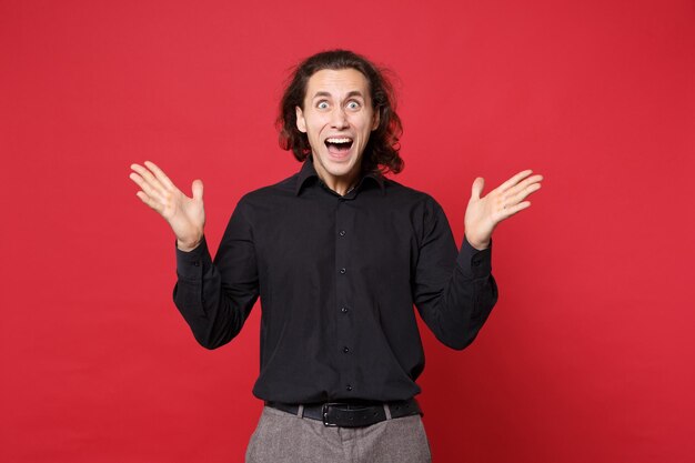 Giovane uomo dai capelli lunghi riccio triste in camicia nera in piedi in posa isolato sul ritratto in studio di sfondo muro rosso. Persone sincere emozioni concetto di stile di vita. Mock up spazio di copia. Problema crisi depressione