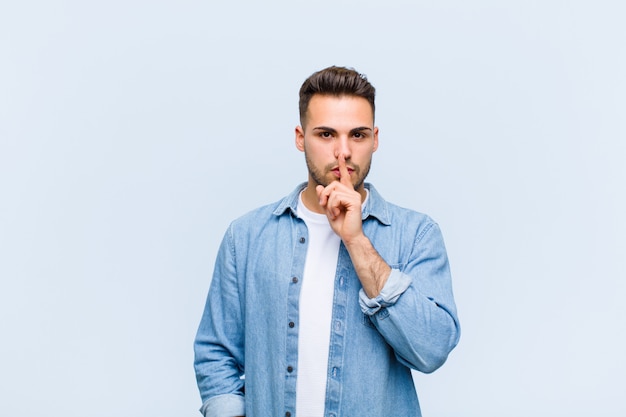 Giovane uomo dall'aspetto serio e incrocio con il dito premuto sulle labbra che richiede silenzio o silenzio, mantenendo un segreto contro il muro blu