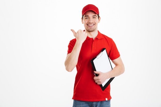 Giovane uomo del corriere in uniforme rossa che sorride e che indica dito da parte sul testo o sul prodotto del copyspace mentre tenendo appunti, isolata sopra spazio bianco