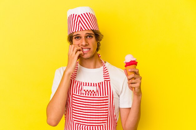 Giovane uomo del gelatiere con il trucco che tiene il gelato isolato su sfondo giallo che si morde le unghie, nervoso e molto ansioso.
