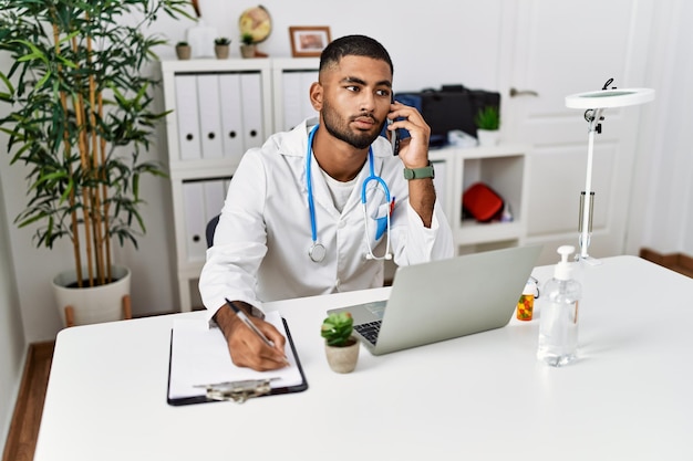 Giovane uomo del Medio Oriente che indossa l'uniforme del medico che parla sullo smartphone in clinica