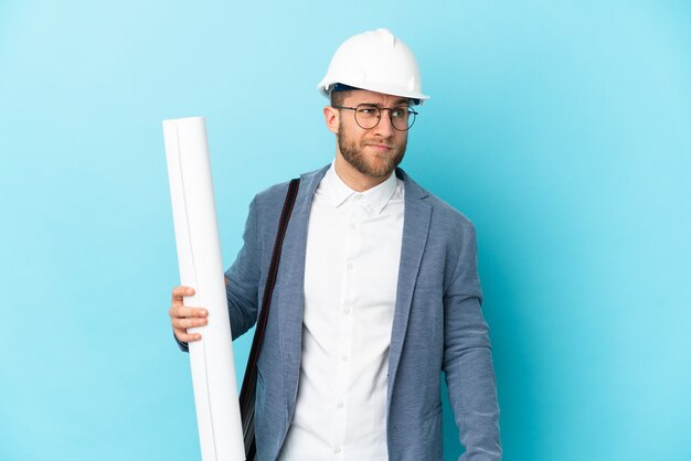 Giovane uomo dell'architetto con il casco e che tiene i modelli sopra fondo isolato che ha dubbi e pensiero