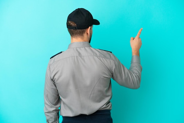 Giovane uomo della sicurezza isolato su sfondo blu che punta indietro con il dito indice