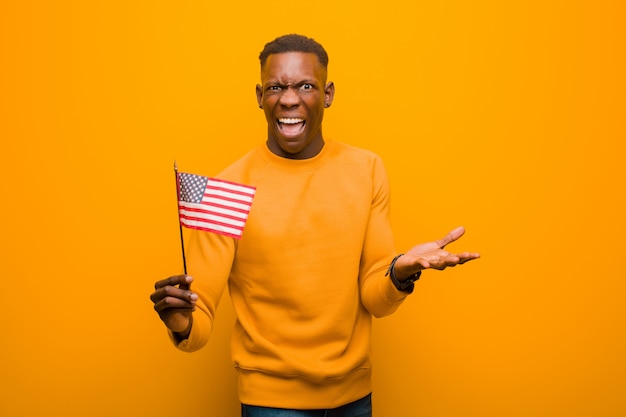 Giovane uomo di colore afroamericano contro la parete arancio che tiene una bandiera degli SUA