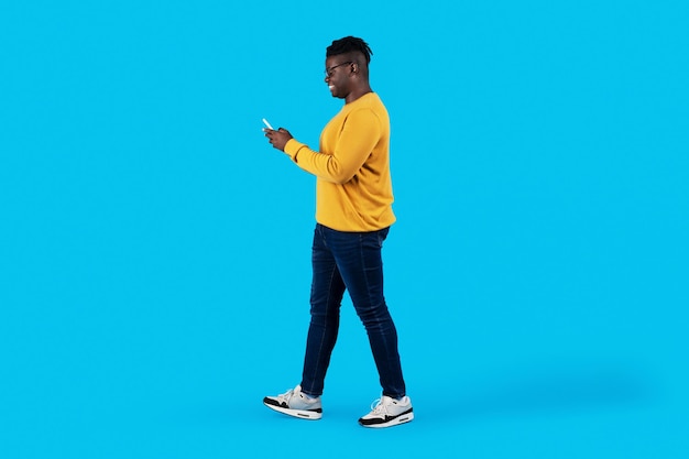 Giovane uomo di colore che cammina con lo smartphone nelle mani su sfondo blu Studio