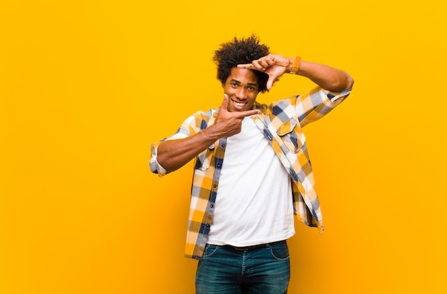 Giovane uomo di colore che ritiene felice sorridere e che fa un ritratto o una cornice con le mani contro la parete arancio