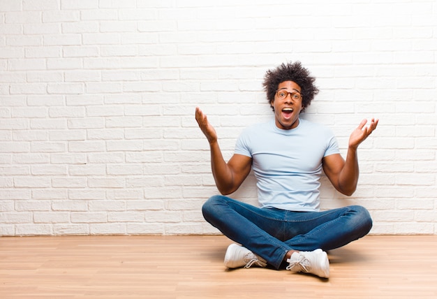 Giovane uomo di colore che si sente felice, eccitato, sorpreso o scioccato, sorridente e stupito di qualcosa di incredibile seduto sul pavimento di casa