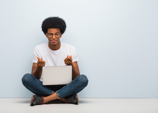 Giovane uomo di colore che si siede sul pavimento con un computer portatile che fa un gesto del bisogno