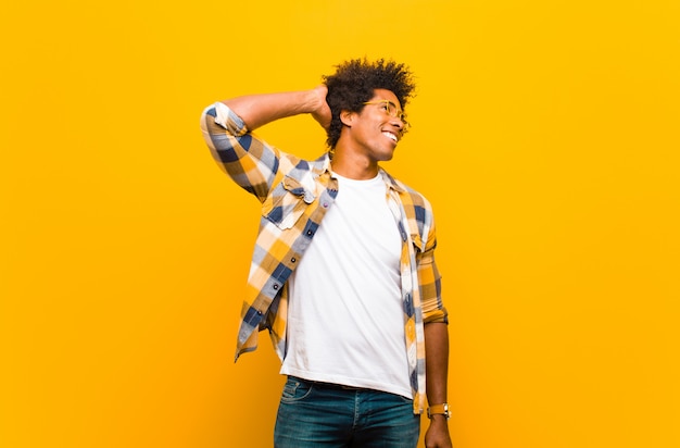 Giovane uomo di colore che sorride allegramente e con disinvoltura, prendendosi per mano a testa con uno sguardo positivo, felice e sicuro contro la parete arancione
