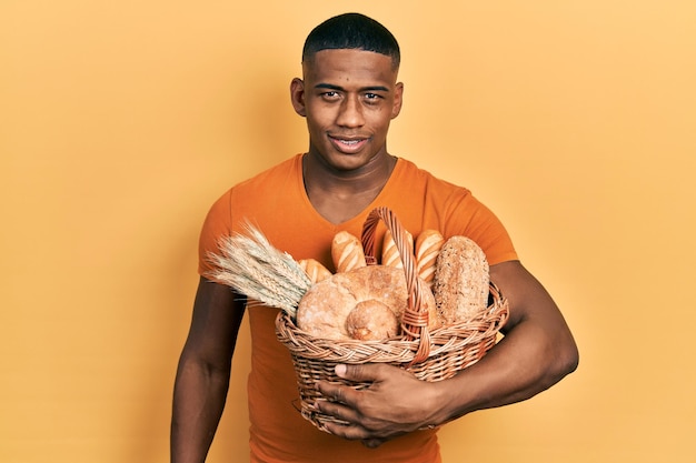 Giovane uomo di colore che tiene cesto di vimini con pane che sembra positivo e felice in piedi e sorridente con un sorriso fiducioso che mostra i denti