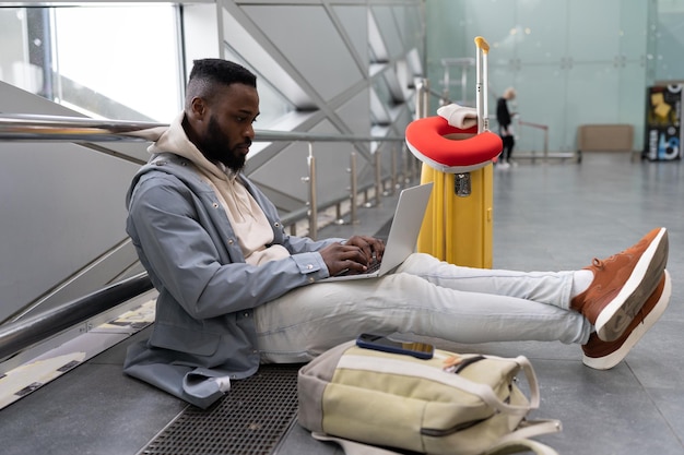 Giovane uomo di colore con il laptop seduto in aeroporto con la valigia che cerca informazioni online sul viaggio