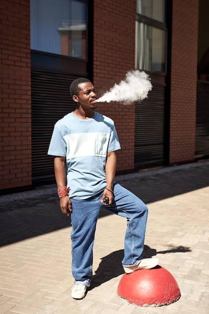 Giovane uomo di colore in maglietta blu e jeans che fumano per strada in una giornata di sole