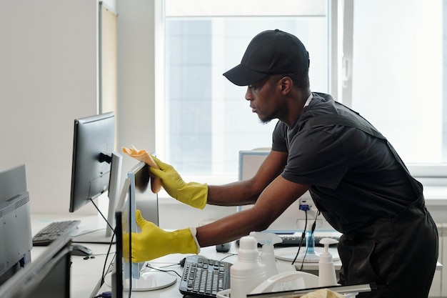 Giovane uomo di colore in uniforme e guanti gialli che puliscono il monitor del computer