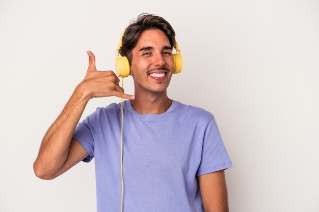 Giovane uomo di razza mista che ascolta musica isolato su sfondo blu che mostra un gesto di chiamata di telefonia mobile con le dita.